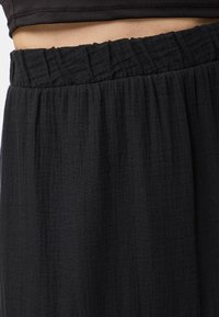 Gros plan sur une ceinture élastique noire sur un pantalon en tissu texturé ample porté avec un top noir court laissant apparaître le ventre.