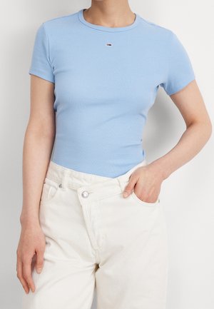 Nainen, jolla on päällään vaaleansininen ribbineulottu lyhythihainen paita ja korkeavyötäröiset off-white-farkut, toinen käsi taskussa valkoista taustaa vasten.