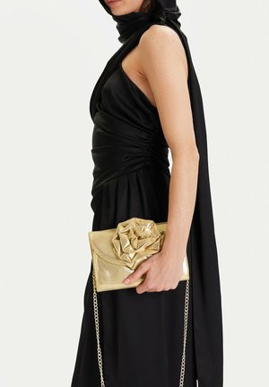 Mujer con vestido negro sin mangas sosteniendo un bolso de mano metálico dorado con un gran detalle de flor y correa de cadena a su lado.