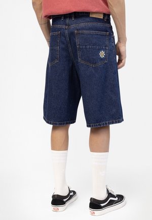 Personne portant un short en denim bleu foncé avec un petit patch marguerite blanche sur la poche arrière, des chaussettes blanches montantes et des baskets noires.