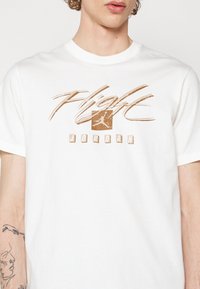 Valkoinen puuvillateepaita, jossa on painettu grafiikka, jossa lukee "Flight" tyylitellyllä ruskealla fontilla ja Jordan-logo neliön sisällä.