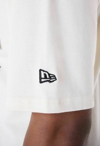 Weißes Baumwoll-T-Shirt mit einer Nahaufnahme eines schwarzen gestickten Logos am Ärmel. Der Stoff wirkt glatt mit einer feinen Textur.