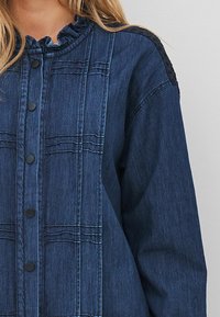 Tamno traper jakna s naboranim ovratnikom, s crnim dugmadi na pritisak i šavovima duž prednje strane i ramena.