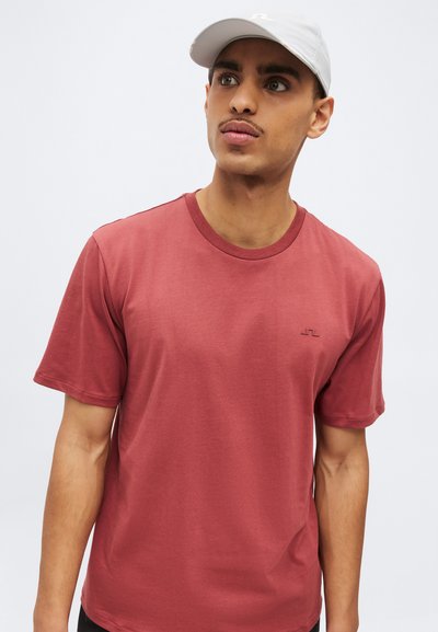 Junger Mann mit hellgrauem Cap und einfarbigem dunkelrotem T-Shirt mit kurzen Ärmeln, der leicht seitlich gegen einen weißen Hintergrund blickt.