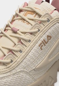 Gros plan d'une basket FILA beige et rose montrant un tissu texturé, les lacets, le logo de la marque sur le côté et une languette rembourrée.