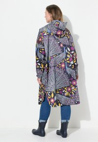 Manteau de pluie à capuche dans un motif floral sombre, avec des couleurs vives comme le violet et le jaune, offrant une coupe ample et des manches longues. Bottes en caoutchouc visibles.