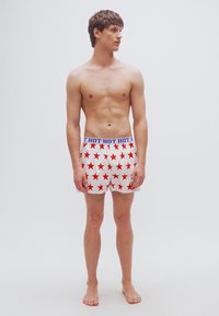 Λευκό μποξεράκι με κόκκινα αστέρια, ελαστική μέση με την επιγραφή "HOT" σε μπλε, που φοριέται από γυμνόστηθο ανδρικό μοντέλο, όρθιο και ξυπόλητο μπροστά σε απλό φόντο.