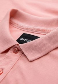 Světle růžová polo tričko s texturovanou tkaninou, knoflíkovou légou a černou nášivkou s nápisem "OMBRE" pobl�íž límce.