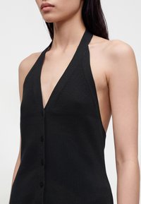 Femme portant un haut noir sans manches à col licou avec des boutons sur le devant, vue du cou à la taille sur un fond uni.