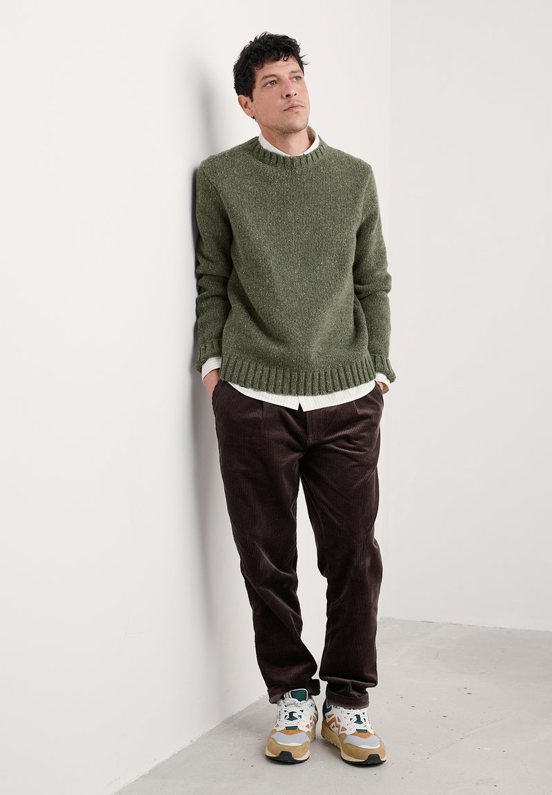 Maglione verde a maglia con orlo e polsini a coste, abbinato a pantaloni in velluto a coste marroni e scarpe da ginnastica multicolori. Design pulito e minimal.