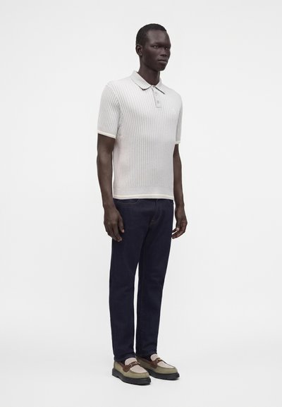 Homme portant un polo en maille gris clair à manches courtes, un jean bleu foncé et des mocassins beiges, debout devant un fond blanc uni.