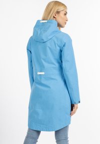 Abrigo impermeable azul claro con capucha, mangas largas, puños abotonados y un detalle ajustado en la parte posterior. Textura suave y diseño hasta la rodilla.