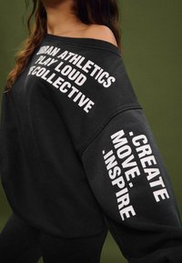 Felpa nera oversize con testo bianco, presenta un design a spalle scoperte e maniche lunghe. Il testo include "Urban Athletics" e "Create Move Inspire."