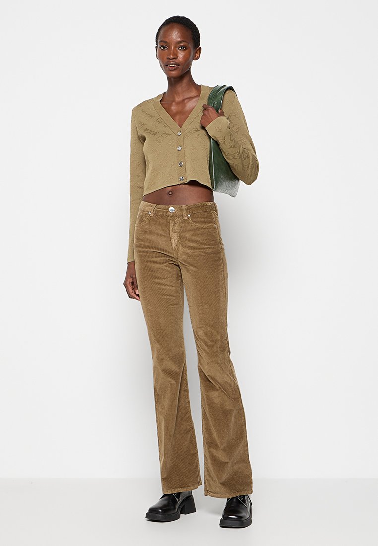 Kardigan w oliwkowym kolorze, krótki, z długim rękawem i zapięciem na guziki, połączony z brązowymi spodniami z korduroju w kształcie dzwonów. Modelka trzyma zieloną torbę, nosi czarne botki.
