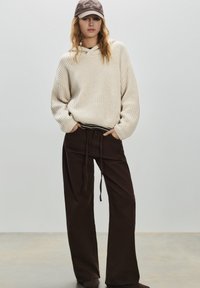 Felpa con cappuccio in maglia color crema con una vestibilità rilassata, abbinata a pantaloni ampi a gamba larga di colore marrone scuro, con vita con coulisse e una texture sottile.