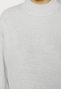 Ljusgrå, stickad tröja med ribbad, hög halsringning och lös passform. Texturen är mjuk med ett fint vävmönster.