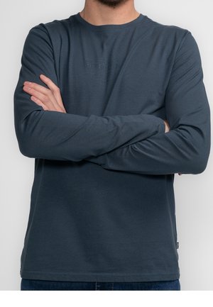 Mężczyzna ze złożonymi ramionami, ubrany w gładką ciemnoniebieską koszulę z długim rękawem i niebieskie dżinsy na jasnym tle.