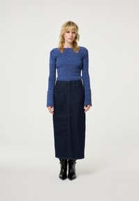 Blauwe gebreide top met lange mouwen, gecombineerd met een donkerdenim midi-rok. Model draagt zwarte puntige enkellaarzen. Eenvoudig ontwerp, getailleerde silhoette.