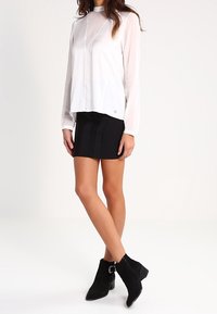 Weiße Langarmbluse mit V-Ausschnitt und transparenter Stoff, kombiniert mit einem schwarzen Minirock. Schwarze Knöchelstiefel mit seitlichem Schnallen-Detail.
