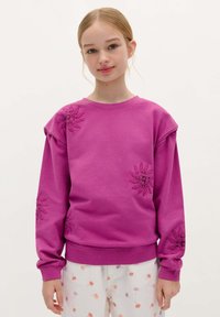 Fuchsia sweatshirt med blomsterbroderi på skuldre og ærmer, ribbede manchetter og kant, båret med hvide bukser med farverige frøblomsttryk.