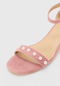 Rosa semskesandal med ankelrem, åpen tå og en bred rem dekorert med hvite falske perler langs fronten. Flat såle.