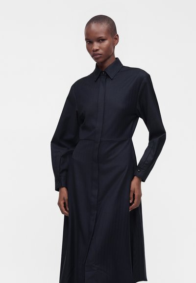 Femme aux cheveux courts portant une robe noire à manches longues boutonnée avec un col, debout devant un fond clair uni.
