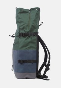 Grön och blå flerpanelryggsäck med rulltoppdesign, framfack med blixtlås, justerbara remmar och svarta metalldetaljer.