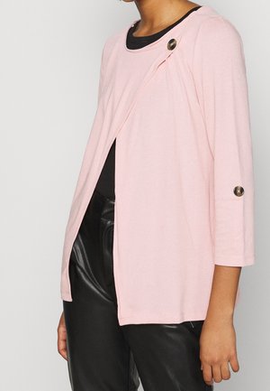 Vrouw die een lichtroze openvallende cardigan draagt met knoopdetails op de schouder en mouw, gecombineerd met een zwarte top en zwarte leren broek.