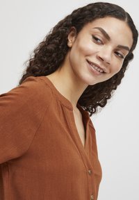 Camicia button-up marrone, con texture, colletto e dettagli delle pieghe. Presenta bottoni in tono oro e una vestibilità rilassata.