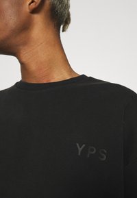 Svart t-shirt i mjukt tyg, med rund halsringning och en subtil "YPS"-logga i en lätt upphöjd textur på bröstet.