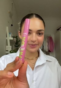 Tube de produit à lèvres rose avec étiquette de texte vert, forme cylindrique, finition brillante, tenu dans une main contre un arrière-plan intérieur flou.