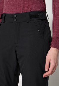 Zwarte broek met een gestructureerde afwerking, voorzien van een brede tailleband, twee knopen en zijzakken. Een rood shirt met lange mouwen is gedeeltelijk zichtbaar.