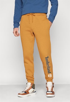 Mężczyzna nosi musztardowe spodnie dresowe Timberland z logo marki na lewej nodze, połączone z białymi, brązowymi i granatowymi wysokimi tenisówkami.