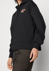 Persoon draagt een zwarte Calvin Klein hoodie met logo op de borst en donkere broek, staand tegen een effen lichte achtergrond.