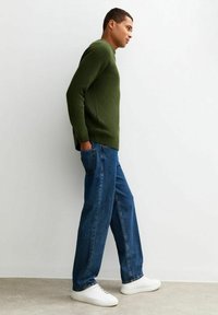 Suéter verde de canalé, jeans azul de corte holgado y zapatillas deportivas blancas. El suéter tiene un dobladillo partido y un escote redondeado. Conjunto casual mostrado de perfil.
