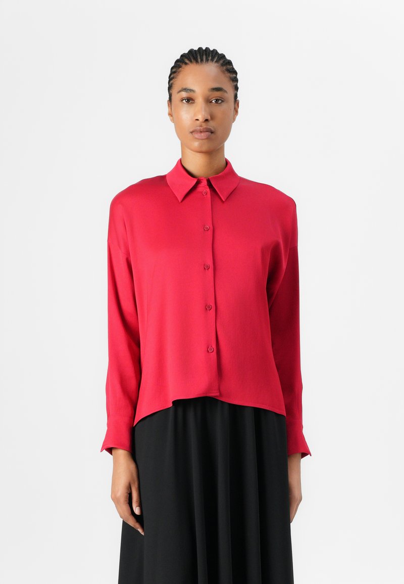 Blouse rouge à manches longues avec un col et une fermeture à boutons, présentant une coupe droite et une texture lisse. Associée à une jupe noire.
