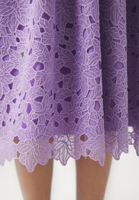 Lila čipkasta krila, ki vsebuje cvetlične vzorce z zapletenimi detajli. Rob je valovit, kar razkriva trdno podlago.