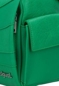 Sac à dos en cuir vert avec une texture lisse, un design structuré et une poche à rabat à l'avant. Comprend un détail de logo argenté sur le côté.