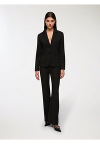 Czarna marynarka dopasowana z dwoma przednimi kieszeniami, połączona z czarnymi spodniami w kształcie dzwonów. Ubranie ma głęboki dekolt w kształcie litery V i eleganckie czarne szpilki.