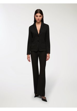 Blazer nero su misura con due tasche anteriori, abbinato a pantaloni neri a campana. L'outfit presenta un profondo scollo a V e tacchi neri eleganti.