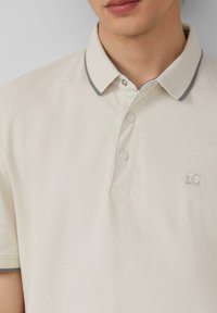 Beige poloshirt met een gestructureerde stof, een kraag met grijze bies en een knoopsluiting met twee knopen, voorzien van een klein logo op de borst.