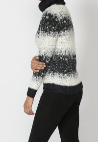 Person, der einen schwarz-weißen, flauschigen Pullover mit Muster und schwarze Hosen trägt, seitlich gezeigt mit einem Arm, der über den Körper gebeugt ist.