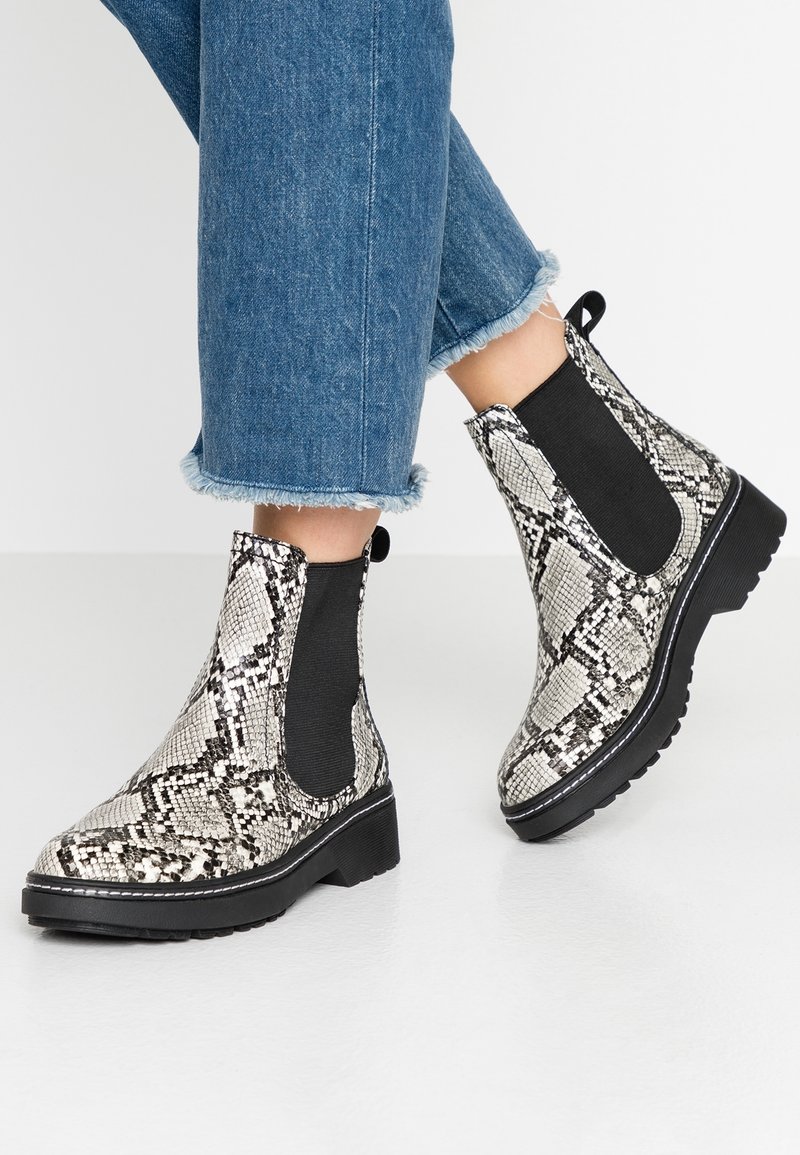 Personne portant des bottines Chelsea à motif peau de serpent montant jusqu'à la cheville, avec des côtés élastiques noirs et des semelles épaisses, associées à un jean bleu à ourlet effiloché.