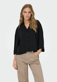 Sort oversized skjorte med knapper og krave, parret med beige bukser med hvide prikker. Blødt stof, afslappet pasform og opbøjede ærmer.