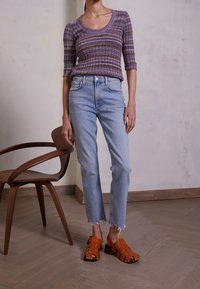 Lila randig stickad tröja med trekvartsärmar, ljusblå högmidjade jeans med fransade fållar, och orangea sandaletter med remmar.