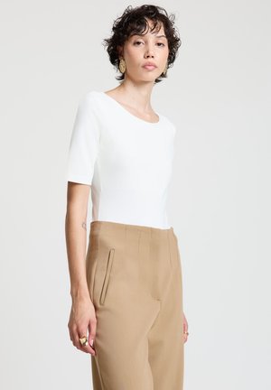 Femme aux cheveux courts et bouclés portant un haut blanc ajusté, un pantalon beige taille haute, des boucles d'oreilles en or et des bagues, sur un fond clair uni.