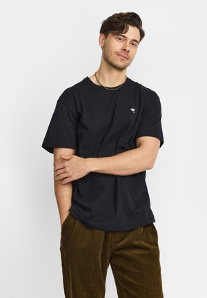 Man met kort bruin haar, dragen een zwart t-shirt en bruine corduroy broek, staand met één hand op zijn andere arm.