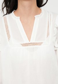 Blouse blanche avec un décolleté en V et un col à volants, ornée de dentelle le long du yoke et confectionnée dans un tissu ample et fluide.