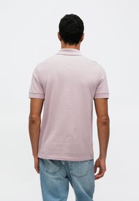 Világos rózsaszín rövid ujjú póló, strukturált gallérral és sima textúrával. Kék farmernadrággal viselve, hátulnézetből.