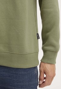 Felpa verde oliva con polsini e fondo a coste; presenta un piccolo etichetta nera sul lato inferiore. Mano visibile appoggiata sul fianco.